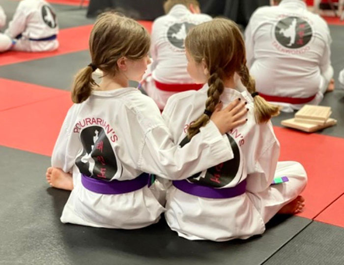 two girls in martial arts uniforms sitting on a mat with their arms around each other's back
