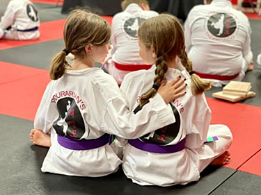 two girls in martial arts uniforms sitting on a mat with their arms around each other's back