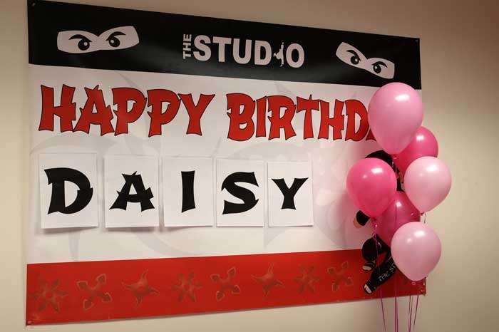 birthday-sign banner that says happy birthday daisy with pink balloons floating in front of it